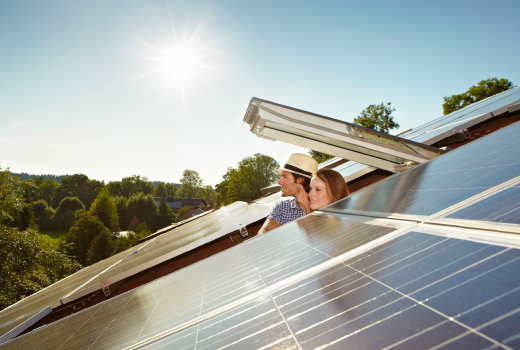 Zwei Personen, schauen aus einem Dachfenster heraus auf ein Dach mit Solarpaneelen. Im Hintergrund sind Bäume und ein klarer Himmel zu sehen.